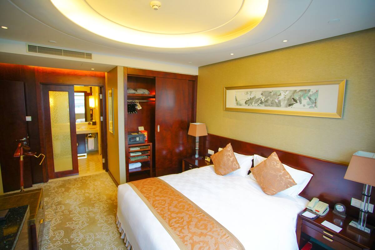 Une chambre d’hôtel avec un grand lit, une table de chevet avec une lampe, une armoire et une salle de bain visible à travers une porte ouverte.