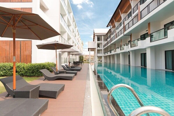 Un área junto a la piscina con sillones de descanso y sombrillas, una piscina y un edificio de varios pisos con balcones.