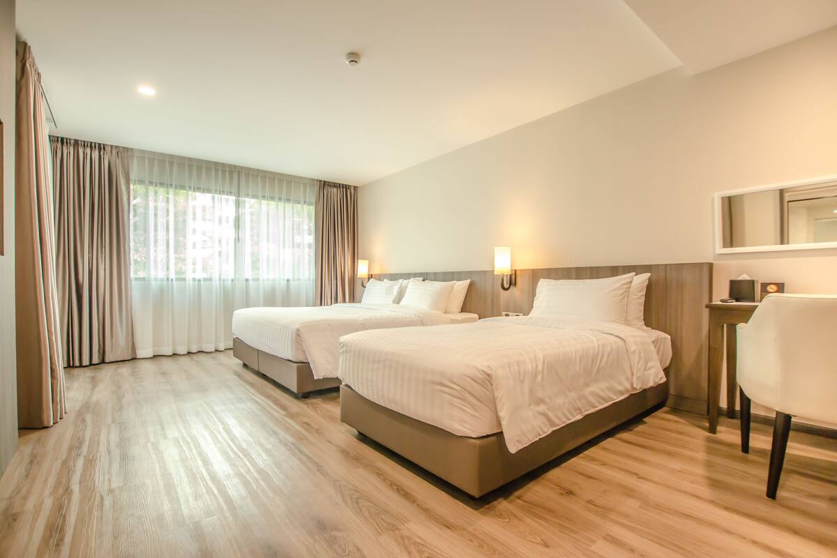 A hotel room with two beds, wooden flooring, a large window with curtains, and a chair.