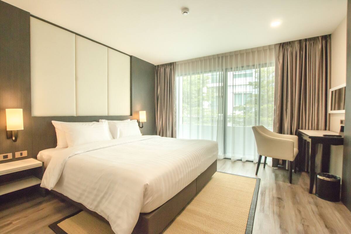 A hotel room with a large bed, a desk, a chair, and a view of the outdoors through the window.