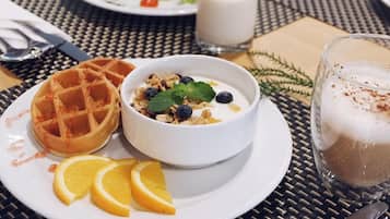 A breakfast setting with a waffle, yogurt with blueberries and granola, orange slices, and a glass of coffee on a woven table.