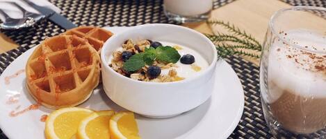 A breakfast setting with a waffle, yogurt with blueberries and granola, orange slices, and a glass of coffee on a woven table.