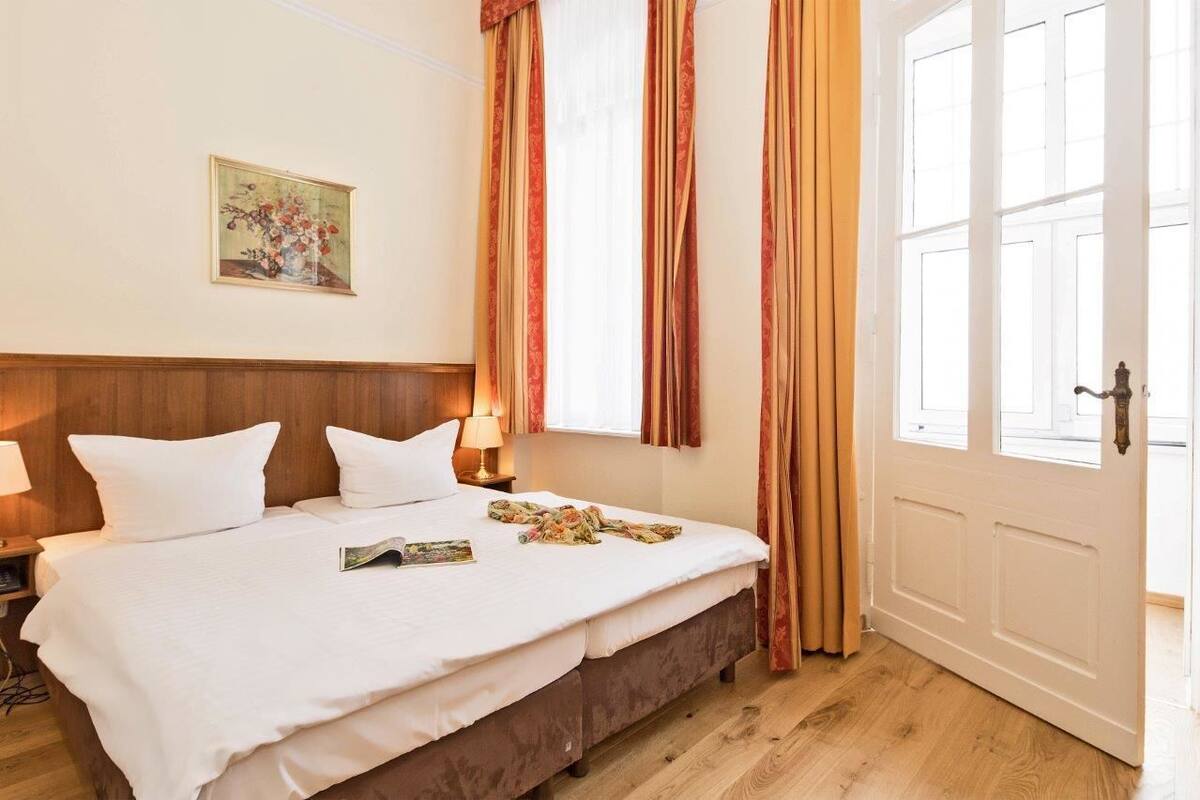 Una habitación de hotel con una cama grande, una mesita de noche, una lámpara, un cuadro en la pared y una ventana con cortinas.