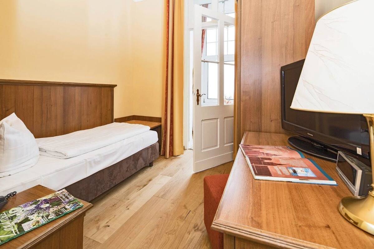 Habitación de hotel con una cama de madera, televisión, escritorio con lámpara y puerta que da a un balcón.