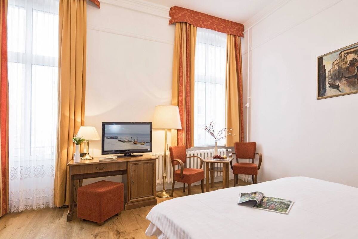 Una habitación de hotel con una cama, un escritorio con una computadora, una silla, una mesita con un florero y cortinas.
