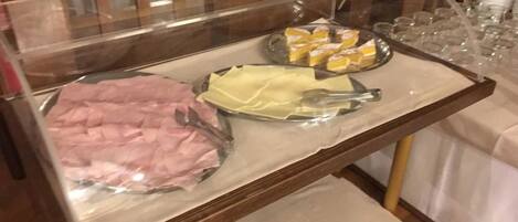 Un carrito de bufé con distintos alimentos, incluyendo un jamón envuelto y un recipiente con elementos amarillos, posiblemente queso o fruta.