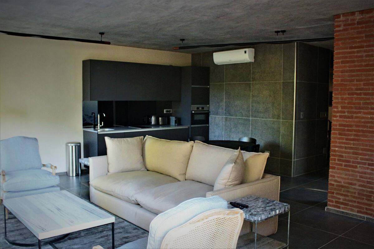 A modern living room with a sofa, a coffee table, and a chair. A kitchen is visible in the background with a microwave and oven.