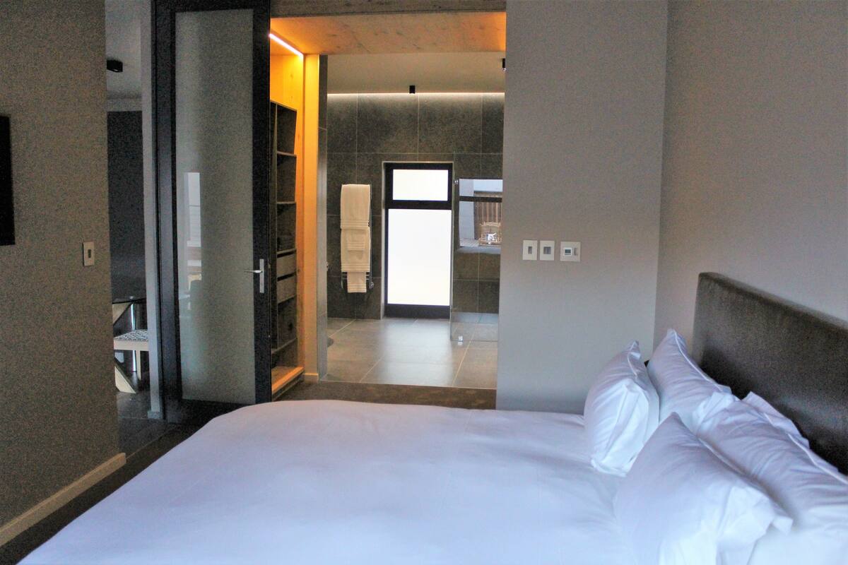 A hotel room with a large bed, a bathroom visible through an open door, and a modern design with neutral colors.