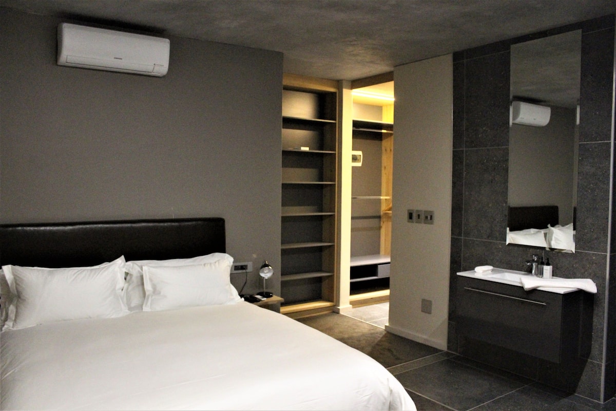 A modern hotel room with a large bed, a walk-in closet, and a bathroom with a mirror and sink.