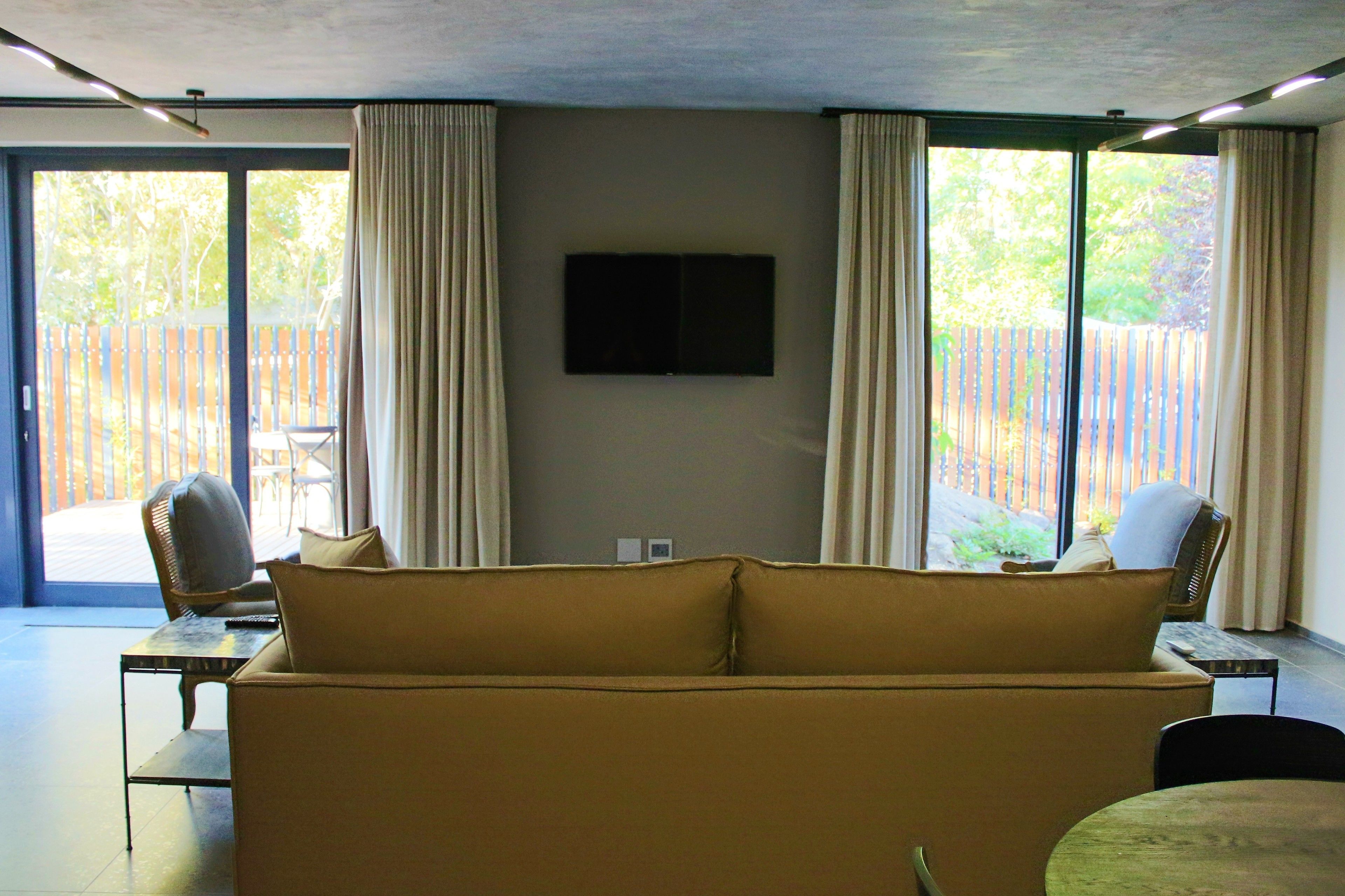A modern living room with a sofa, two chairs, a TV mounted on the wall, and large windows with curtains.