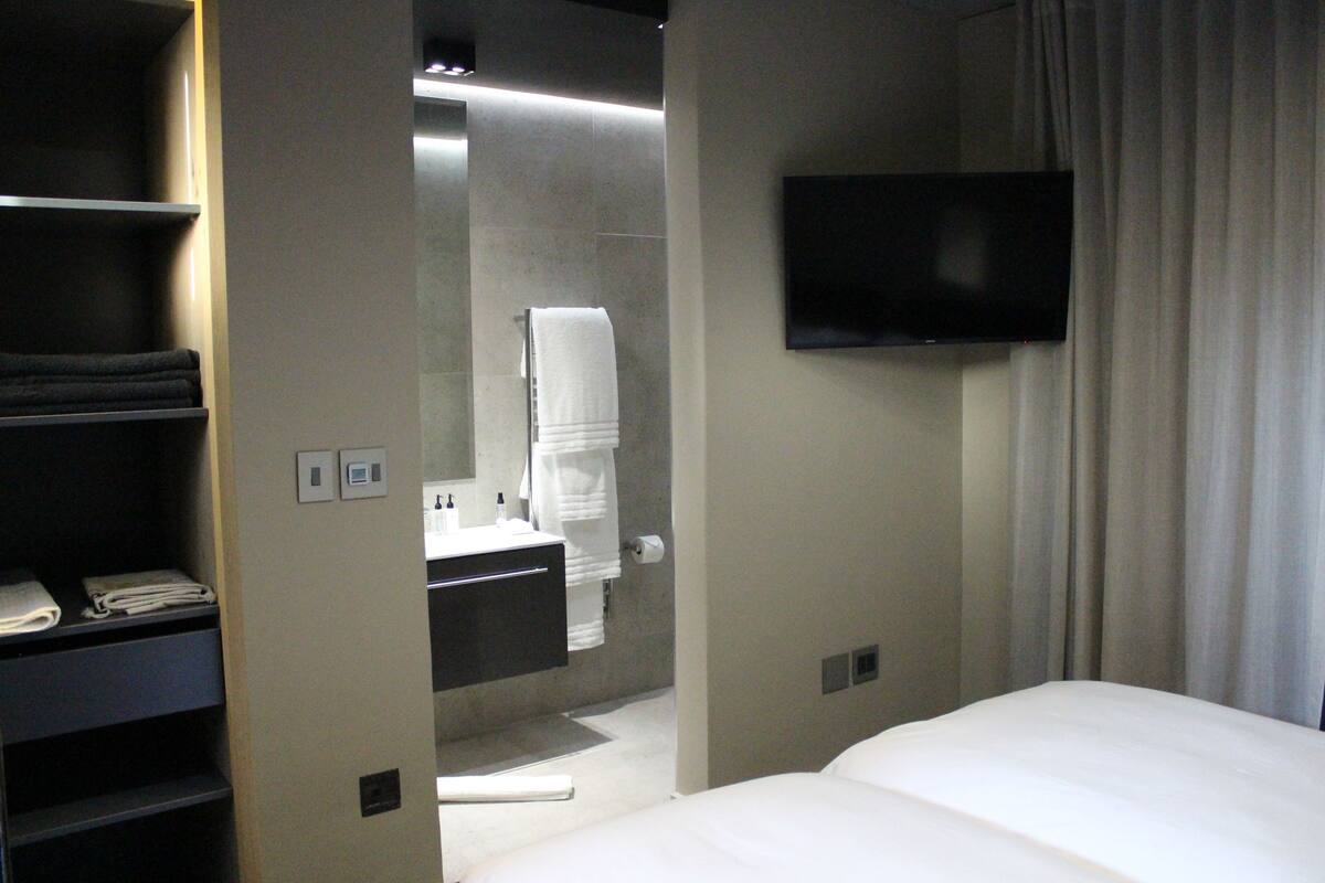 A hotel room with a bed, a TV mounted on the wall, a bathroom with a mirror and towel rack, and a shelf with folded towels.