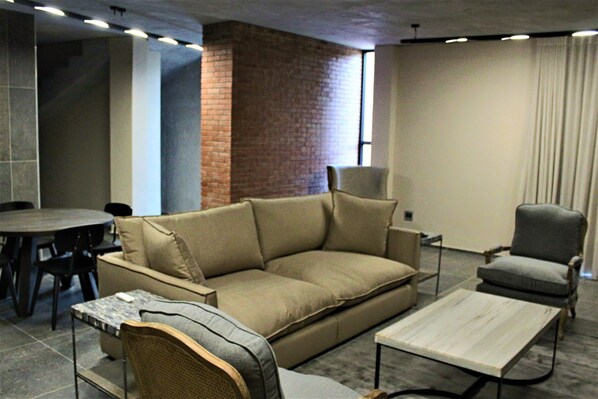 A modern living room with a beige sofa, a grey armchair, a small wooden coffee table, and a dining area with a round table and chairs.