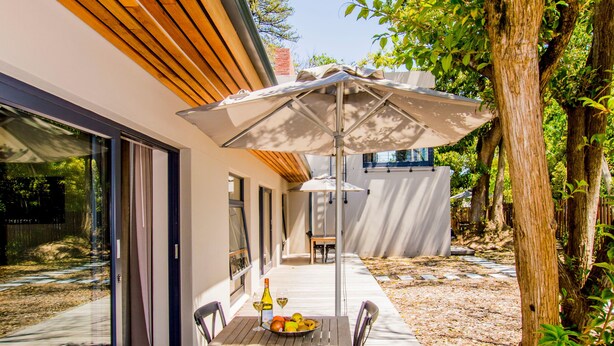 Une terrasse en bois avec une table et des chaises, une porte vitrée donnant sur un bâtiment moderne, et un arbre en arrière-plan.