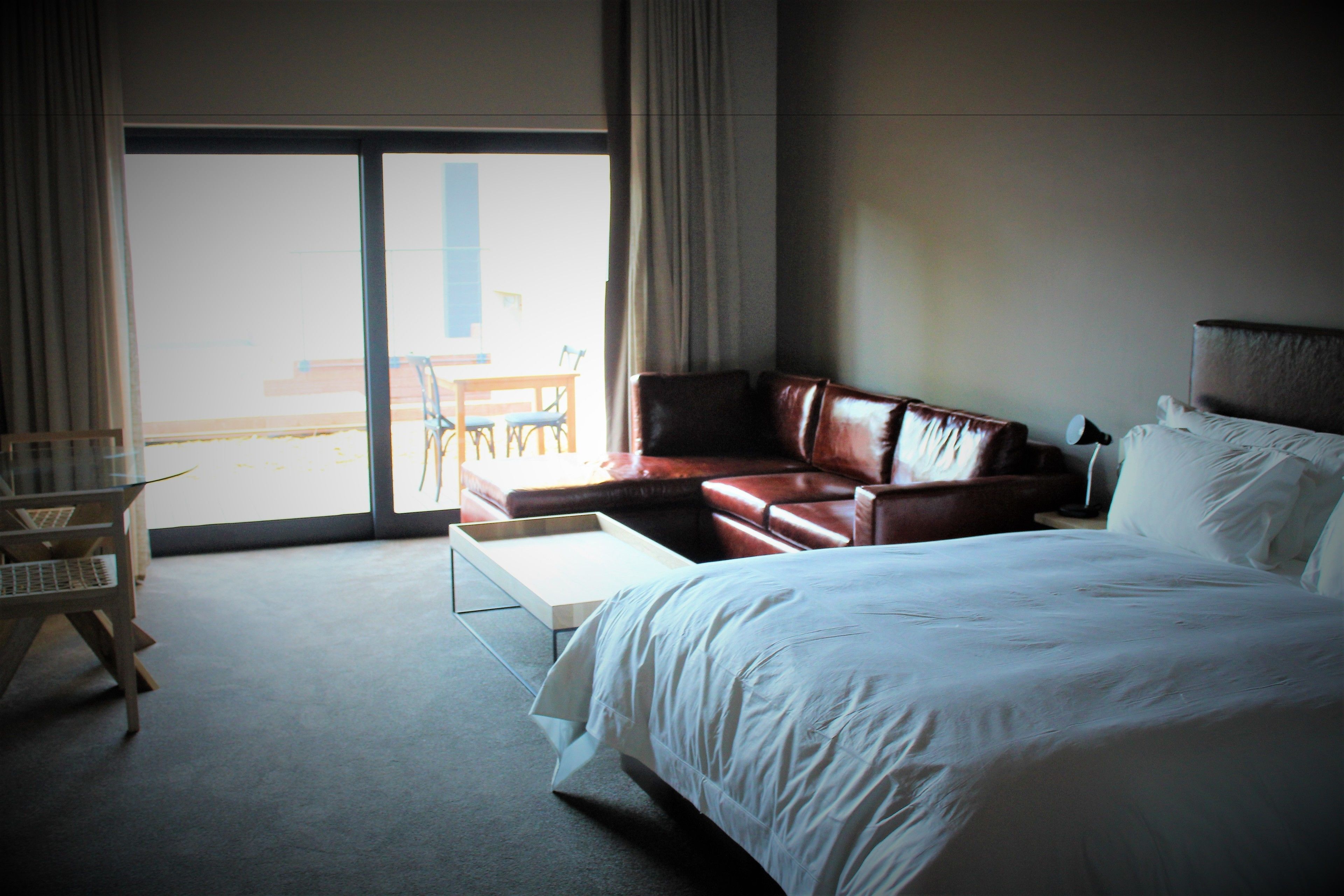 Ein Hotelzimmer mit einem großen Bett, einer Ledersofa, einem kleinen Tisch und Blick ins Freie durch eine Schiebetür.
