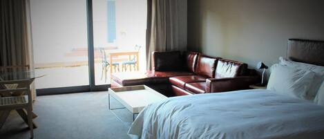 Ein Hotelzimmer mit einem großen Bett, einer Ledersofa, einem kleinen Tisch und Blick ins Freie durch eine Schiebetür.