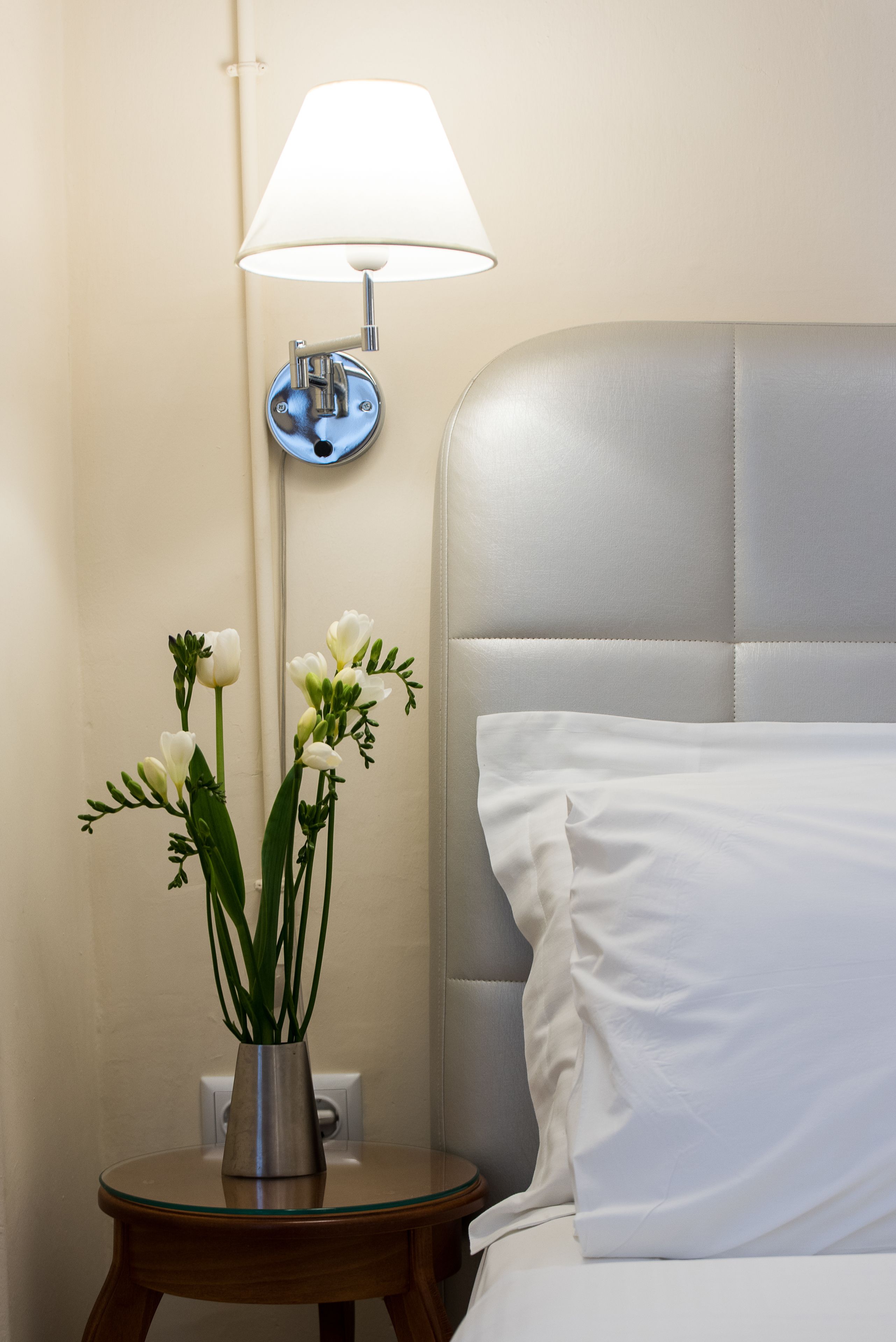 A neatly made bed with a white pillow and a headboard, a bedside table with a lamp and a vase of flowers.