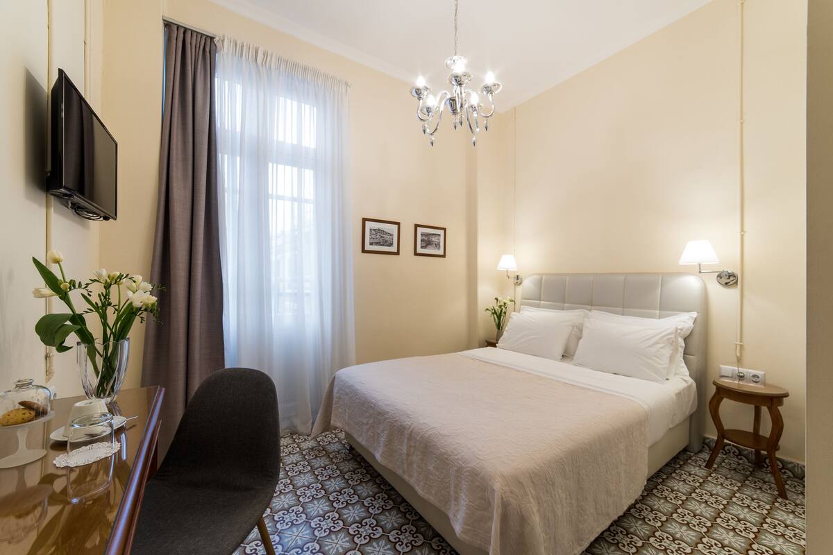Habitación de hotel con cama, televisor, candelabro, escritorio con silla y ventana con cortinas.