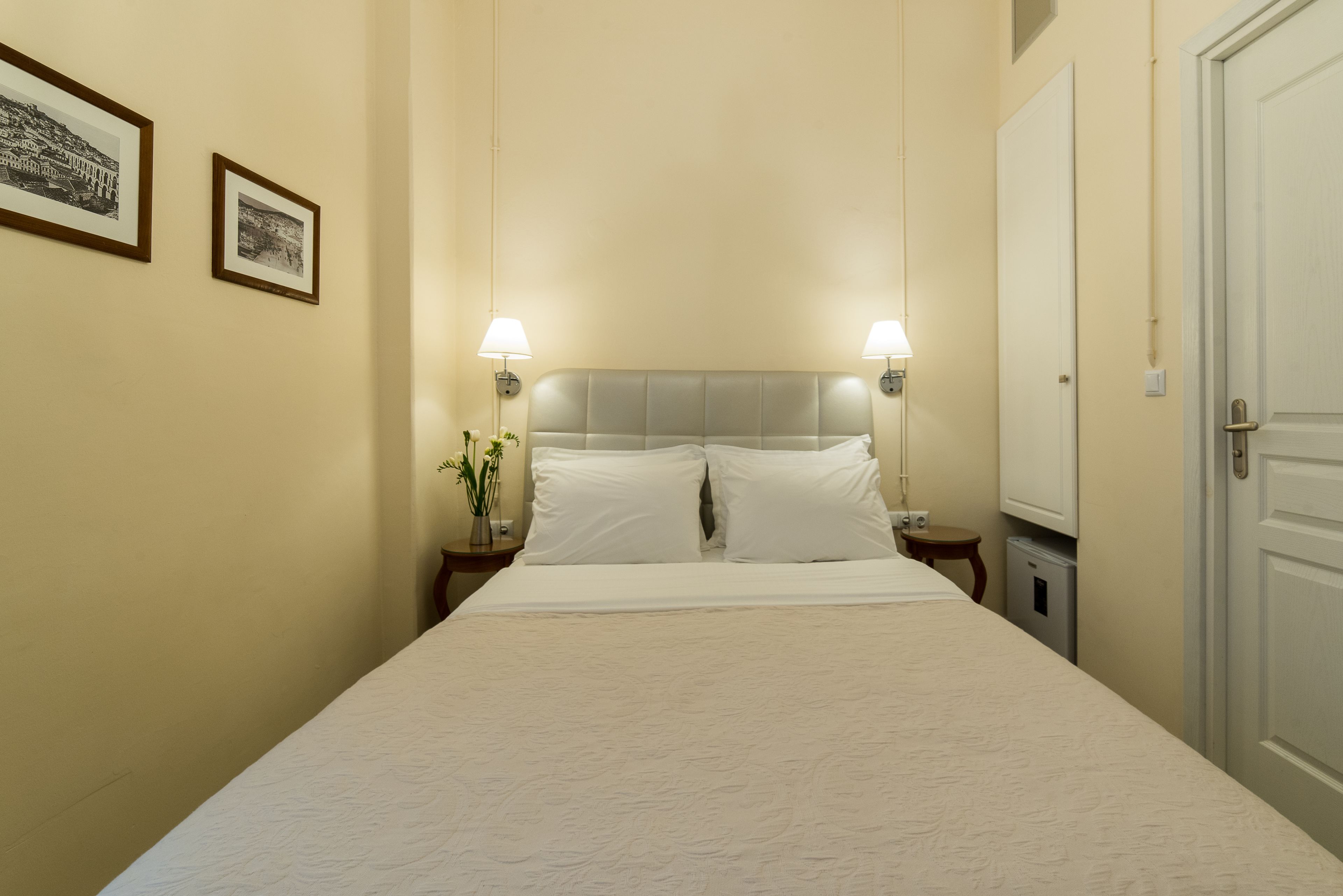 Een netjes opgemaakt bed met witte lakens, twee bedlampen, een nachtkastje met een bloevas en een deur aan de rechterkant.