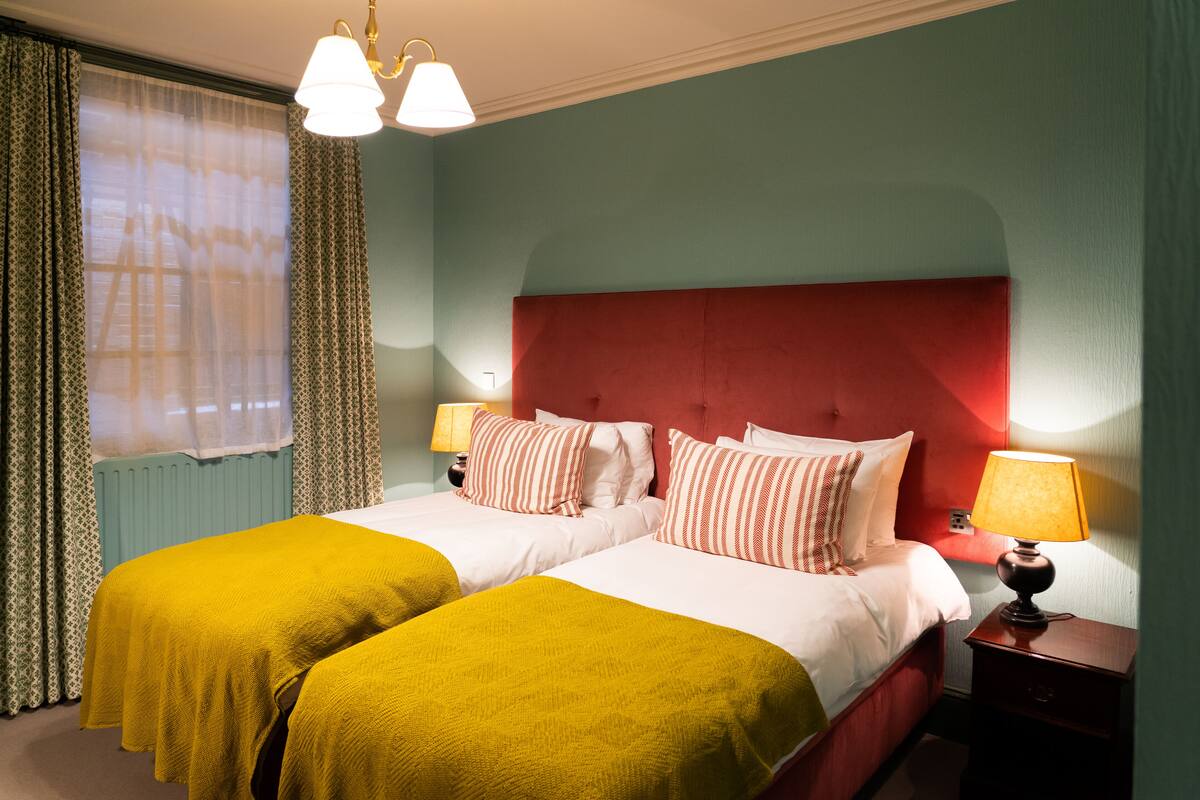 Habitación de hotel con dos camas, cabecera roja y mesita de noche con lámpara.