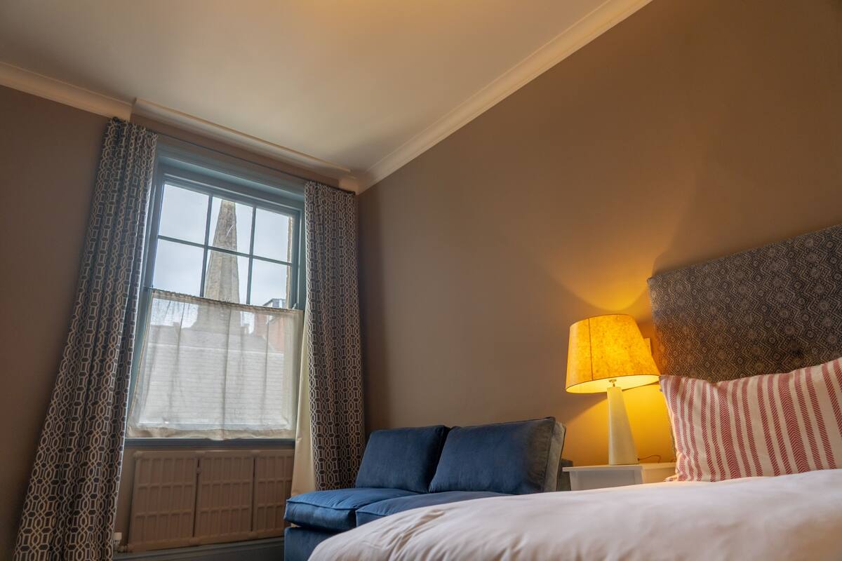 Una habitación de hotel con cama, mesita de noche con lámpara, ventana con cortinas estampadas y radiador.