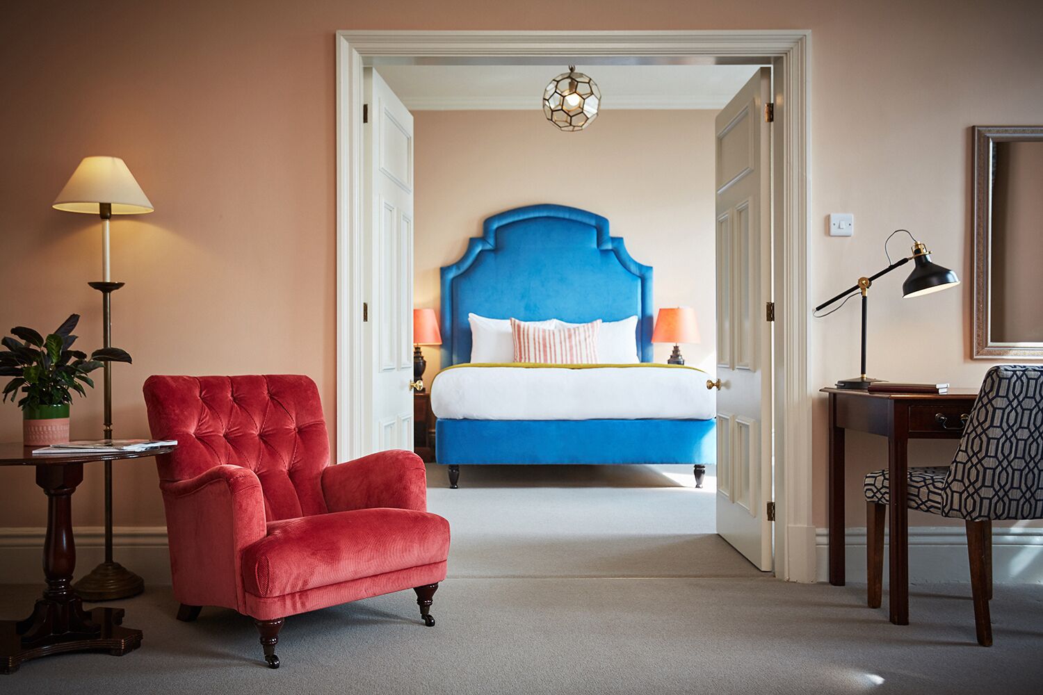 Ein Hotelzimmer mit blauem Stoffkopfende, einem roten Samt-Sessel, einem Schreibtisch mit Lampe und einem Blumentopf.
