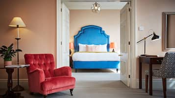 A hotel room with a blue upholstered headboard, a red velvet armchair, a desk with a lamp, and a potted plant.
