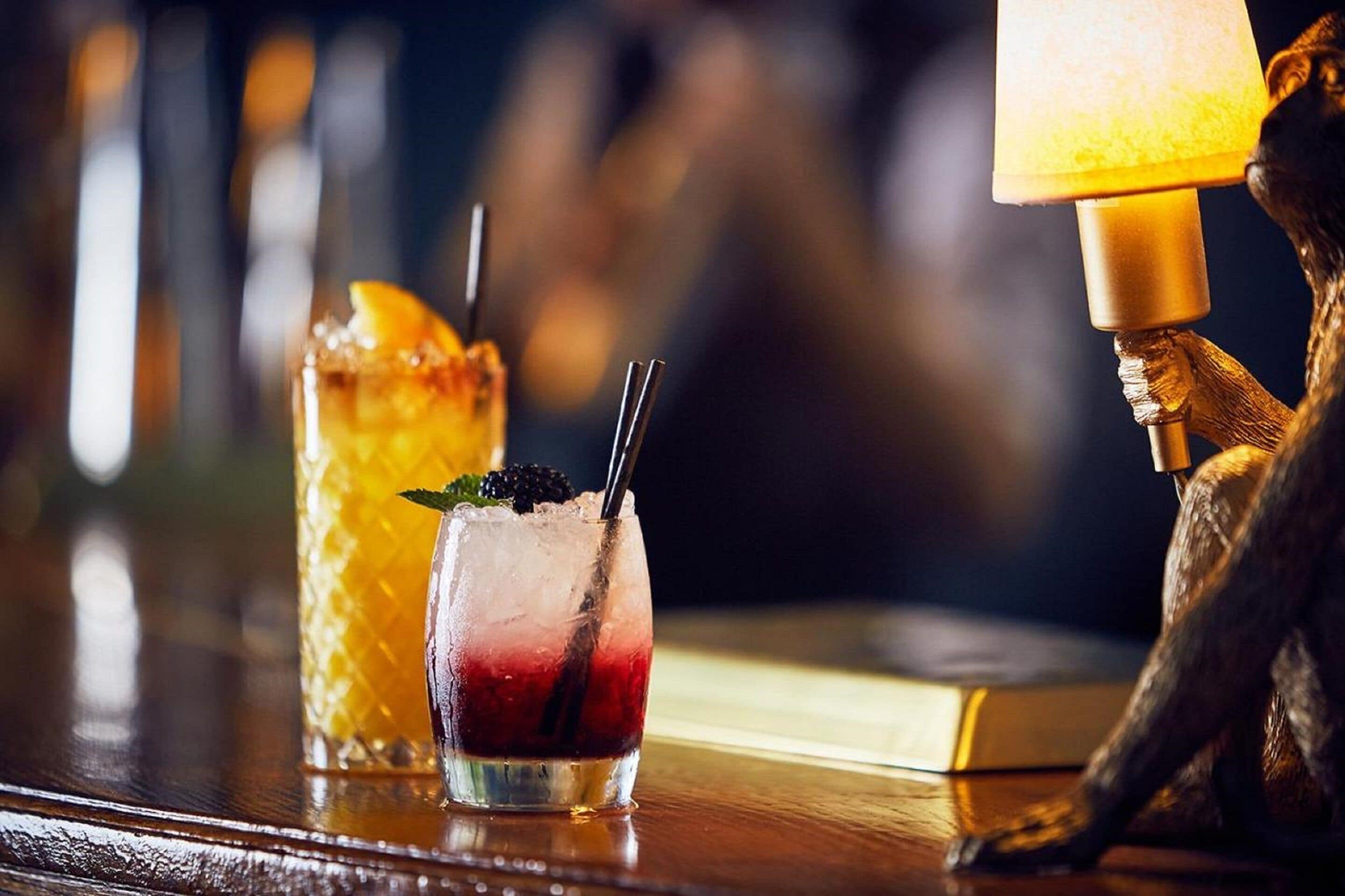 Two drinks on a bar counter with a lamp and a statue in the background.