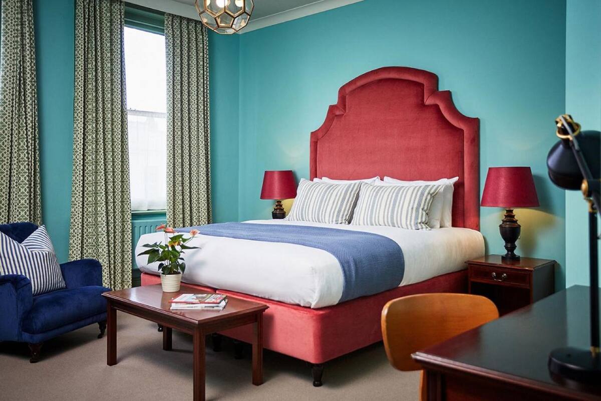 Un dormitorio con cabecera tapizada en rojo, una cama con ropa de cama a rayas, dos mesitas de noche con lámparas, un sillón azul y una mesa de centro de madera.