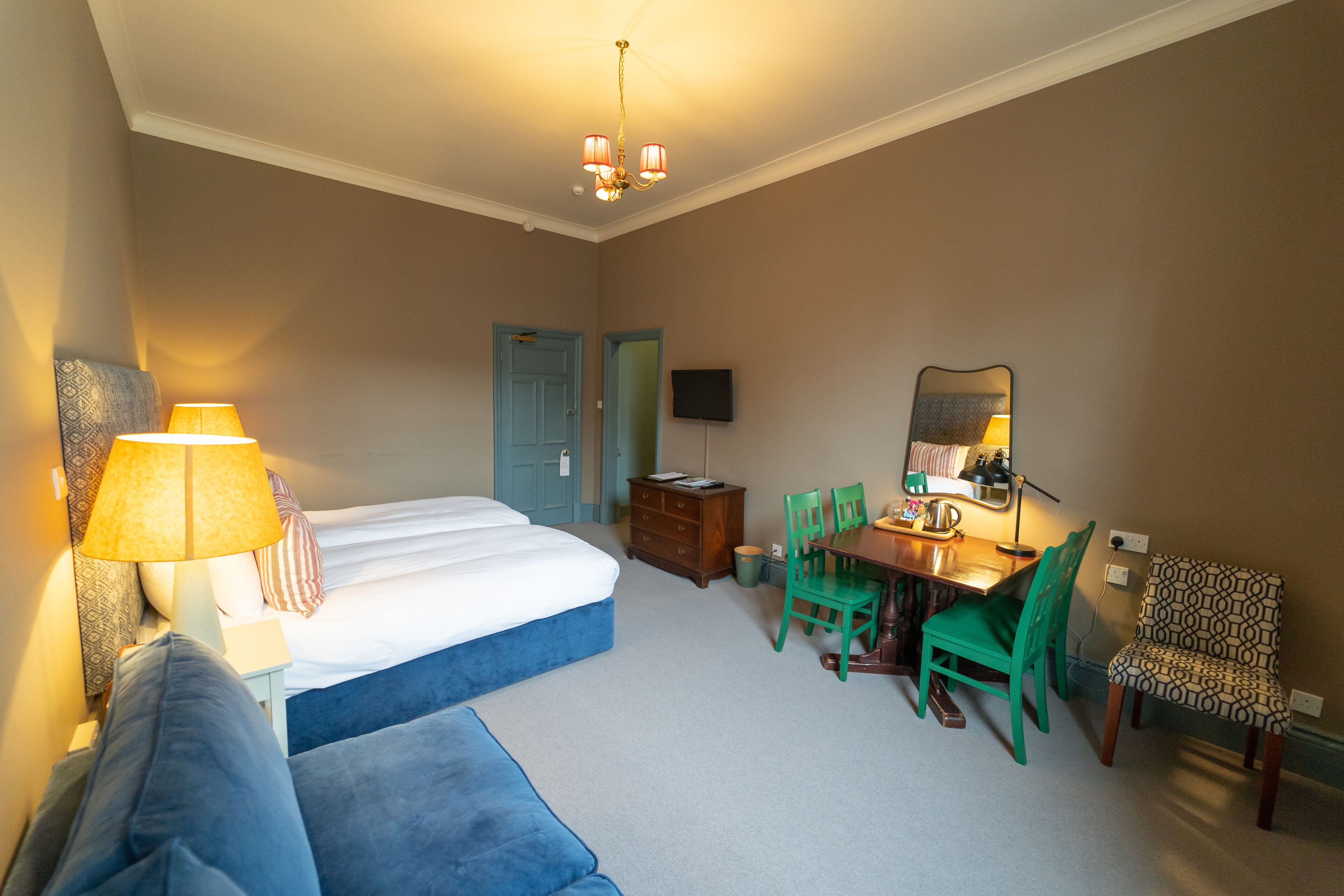 Quarto de hotel com uma cama, escrivaninha e cadeiras, televisão e luminária.