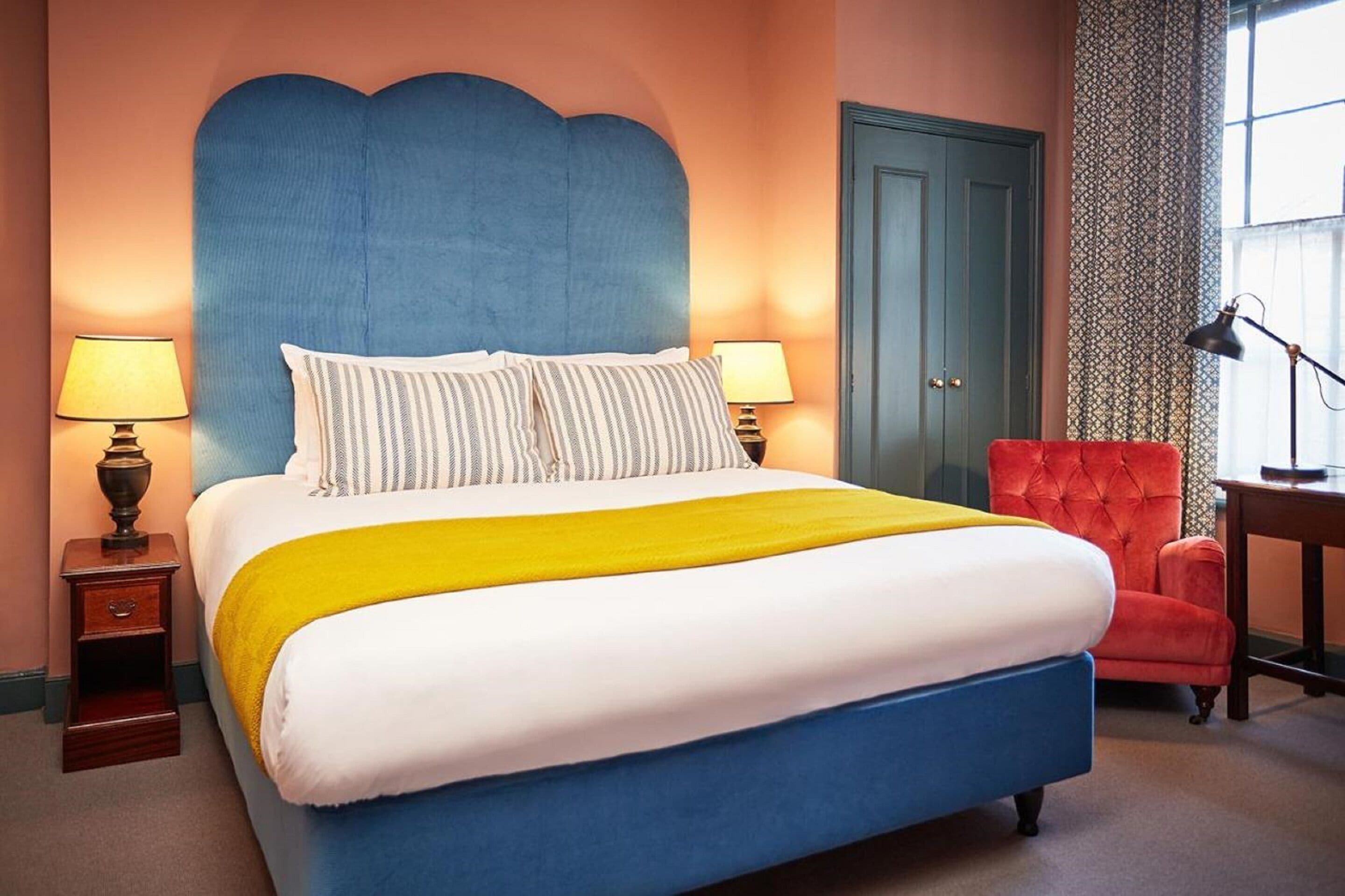 A bedroom with a large bed, two bedside lamps, a red armchair, and a desk with a lamp.