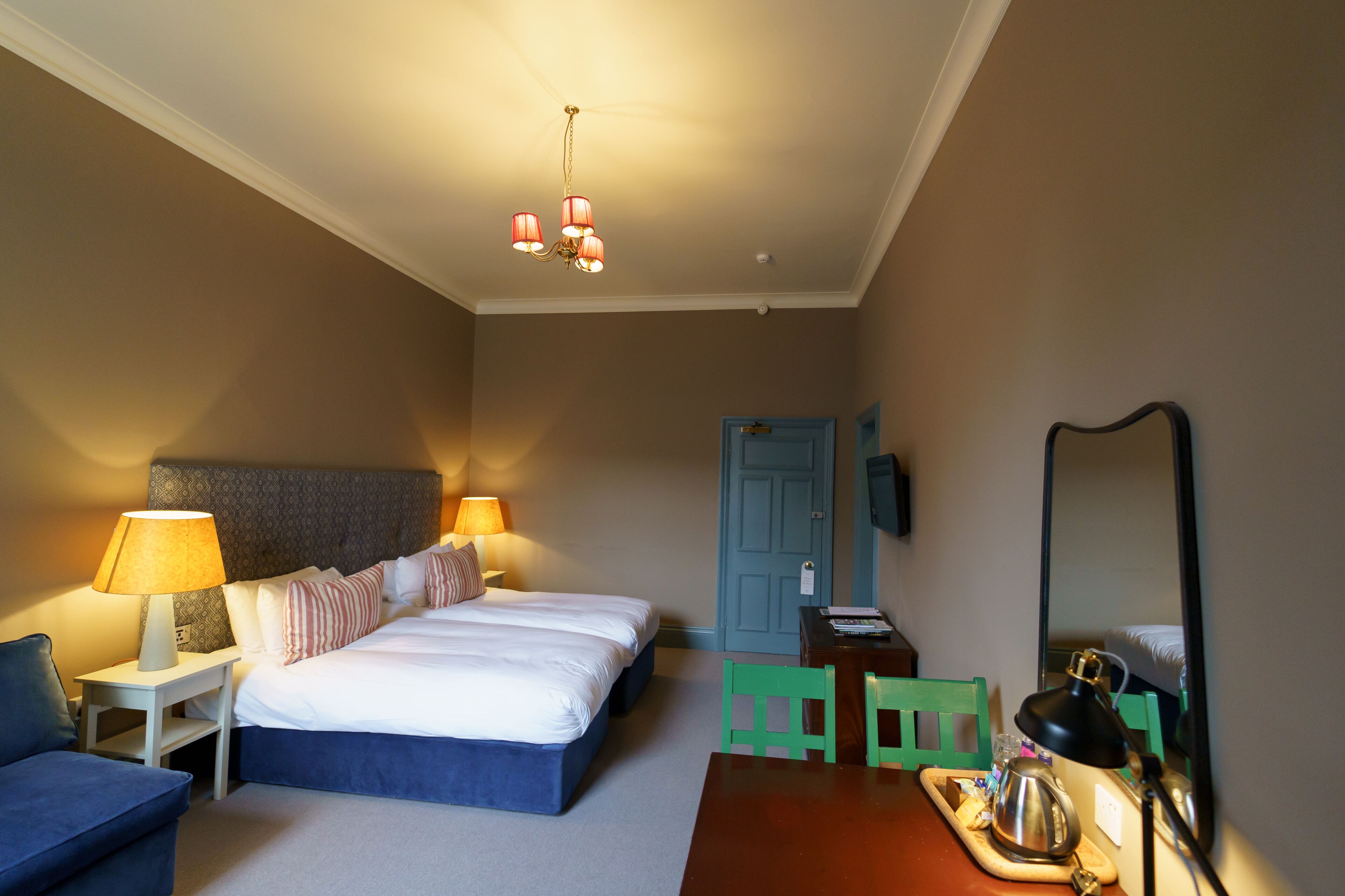 A hotel room with a bed, two bedside tables with lamps, a desk with a chair, a mirror, and a door.
