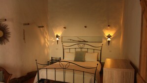A bedroom with a metal-framed bed, two wall-mounted lamps, a wooden nightstand, and a wicker chair. - Is Scabas (Solarussa)