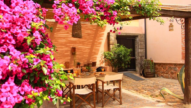 An outdoor patio with a table and chairs, surrounded by vibrant pink flowers and potted plants. - Is Scabas (Solarussa)