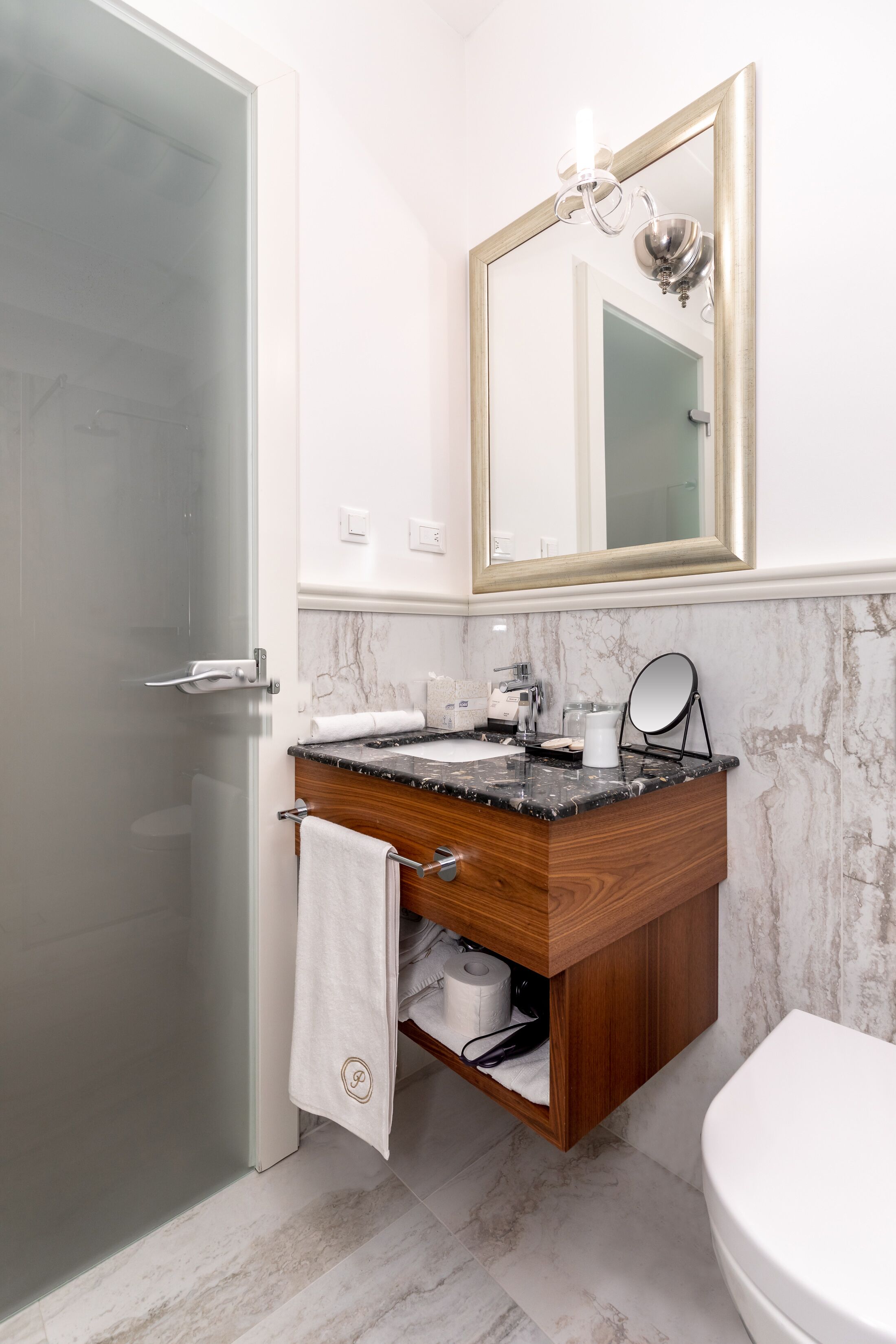Un baño con mampara de vidrio, lavamanos de mármol, tocador de madera, inodoro blanco y espejo con marco dorado.