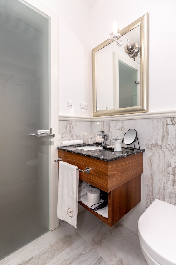 Un baño con mampara de vidrio, lavamanos de mármol, tocador de madera, inodoro blanco y espejo con marco dorado.