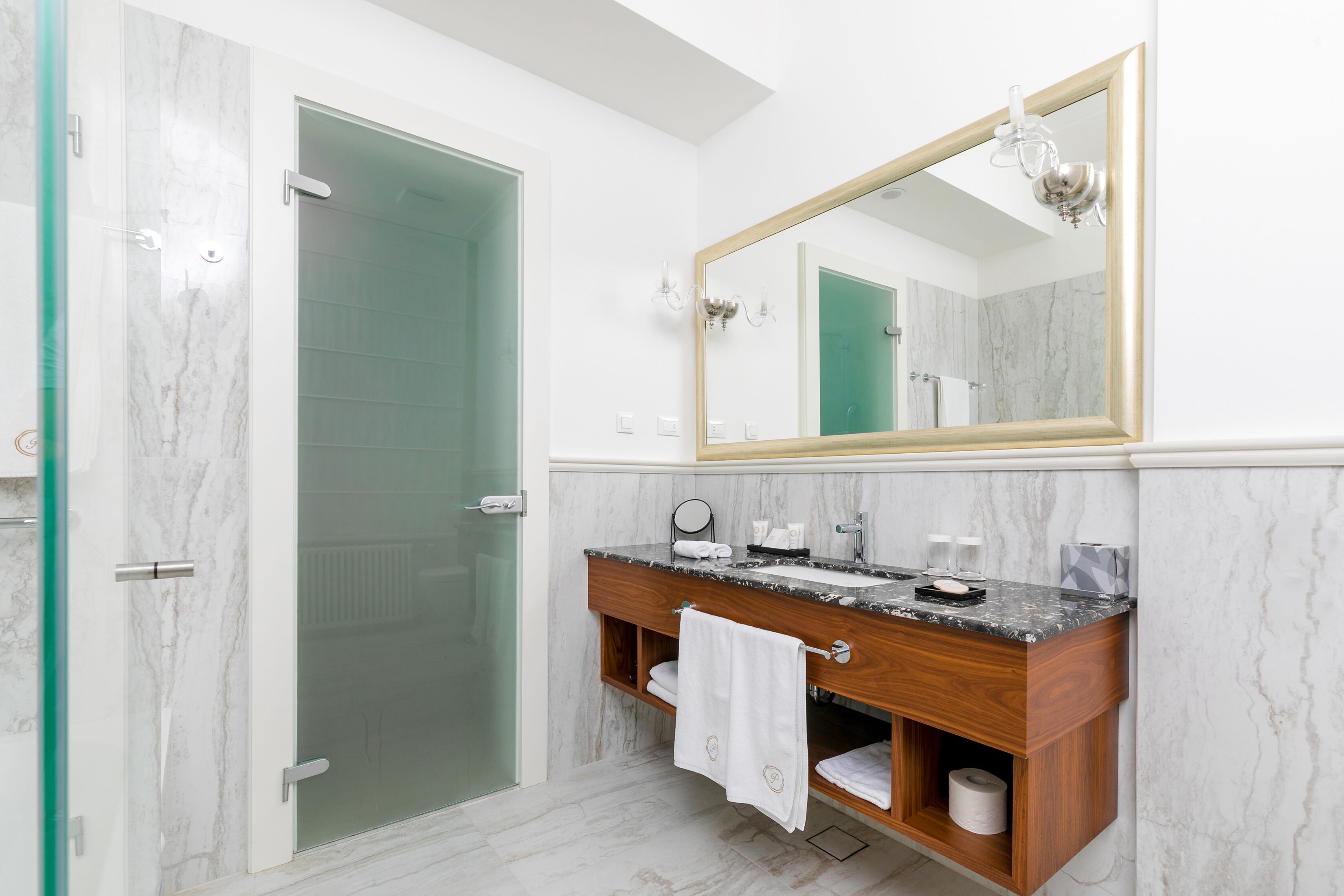 A bathroom with a glass shower door, a wooden vanity with a marble countertop, a large mirror, and a towel rack.