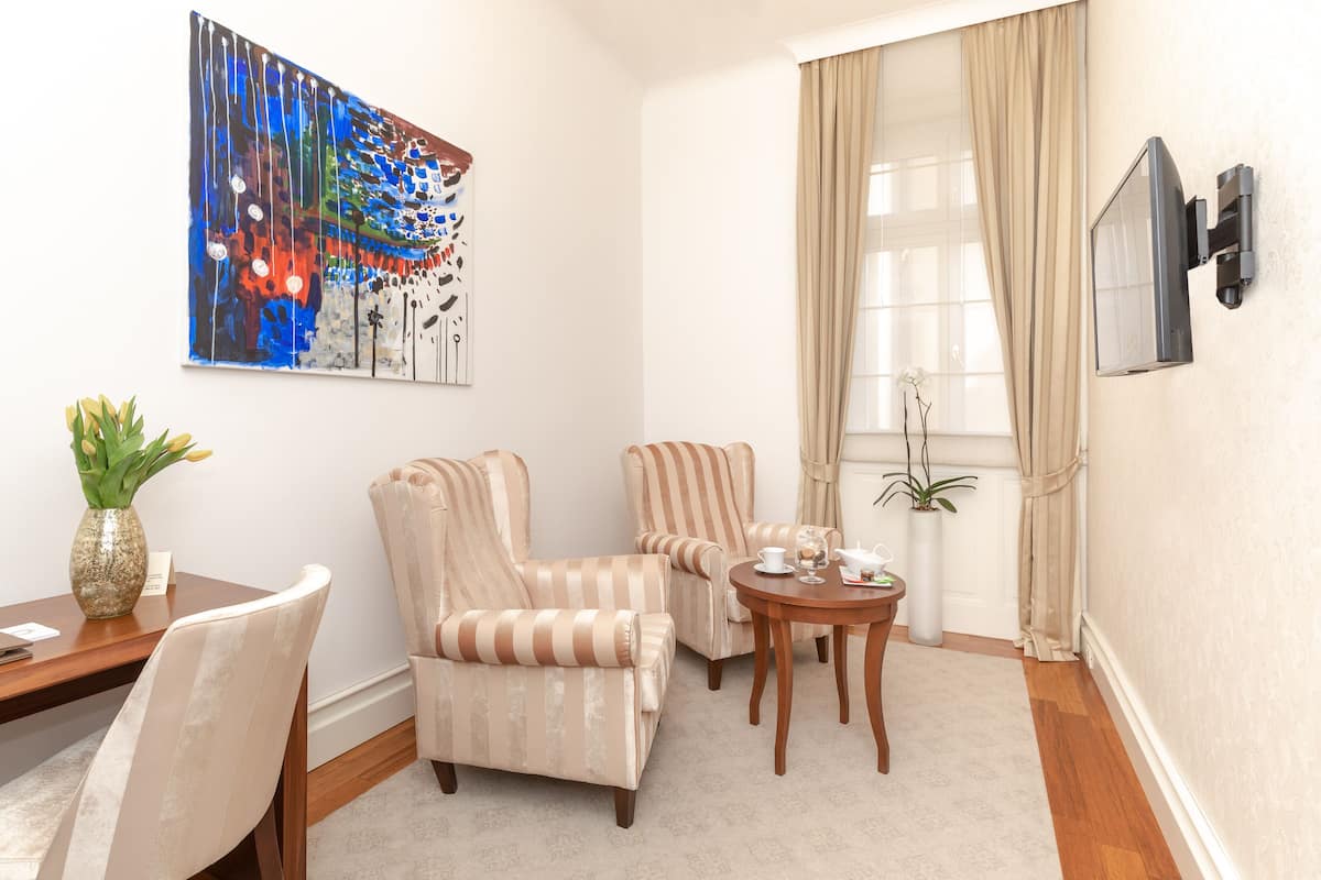 Een kamer met gestreepte fauteuils, een klein rond tafeltje, een schilderij, een aan de muur bevestigde televisie en een raam met gordijnen.