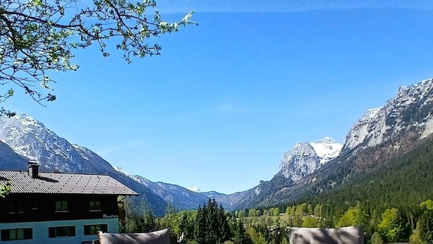 A mountain view with a building and two wicker chairs with a table in the foreground. - Ferienwohnung Hochkalter, 1-2 Erwachsene + 1 Kind, 40 qm, Dusche/WC, TV, W-LAN, Ostbalkon, 1 Schlafzimmer, Nichtraucher (Ramsau bei Berchtesgaden)