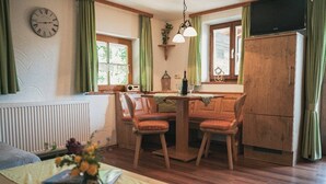A cozy room with a wooden dining table, two chairs, a clock, and a television on a cabinet. - Ferienwohnung Hochkalter, 1-2 Erwachsene + 1 Kind, 40 qm, Dusche/WC, TV, W-LAN, Ostbalkon, 1 Schlafzimmer, Nichtraucher (Ramsau bei Berchtesgaden)