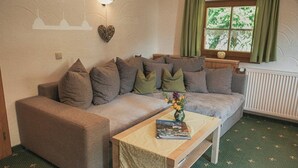 A cozy living room with a sofa, a coffee table, a wall-mounted light, and a window with curtains. - Ferienwohnung Hochkalter, 1-2 Erwachsene + 1 Kind, 40 qm, Dusche/WC, TV, W-LAN, Ostbalkon, 1 Schlafzimmer, Nichtraucher (Ramsau bei Berchtesgaden)