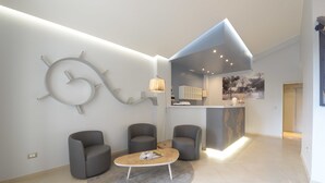 Un interior moderno con un mostrador de recepción, dos sillones grises, una mesa baja con revistas y una gran obra de arte abstracta en la pared.