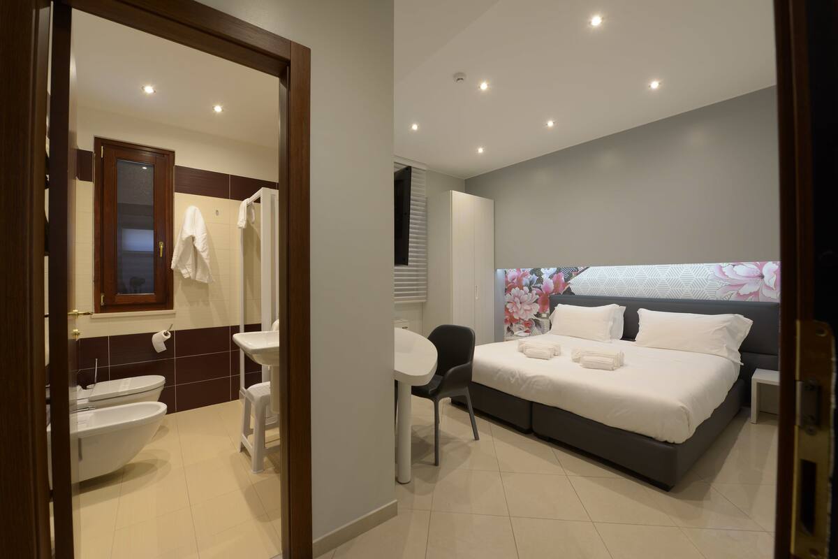 Ein modernes Hotelzimmer mit einem großen Bett, einem Badezimmer mit Toilette und Waschbecken sowie einem kleinen Tisch mit Stuhl.