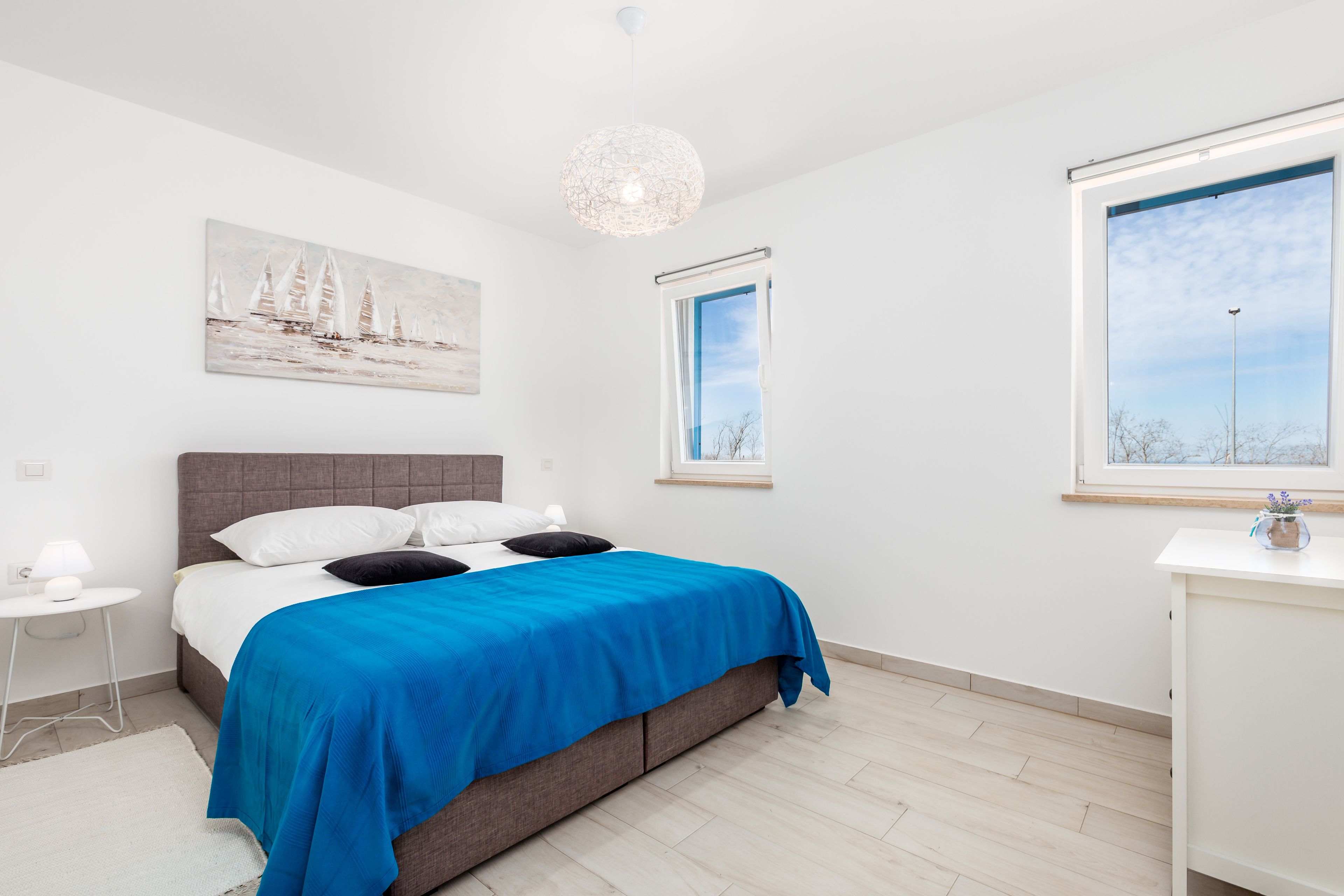 A bedroom with a bed, a nightstand, a desk, a window, and a painting on the wall.