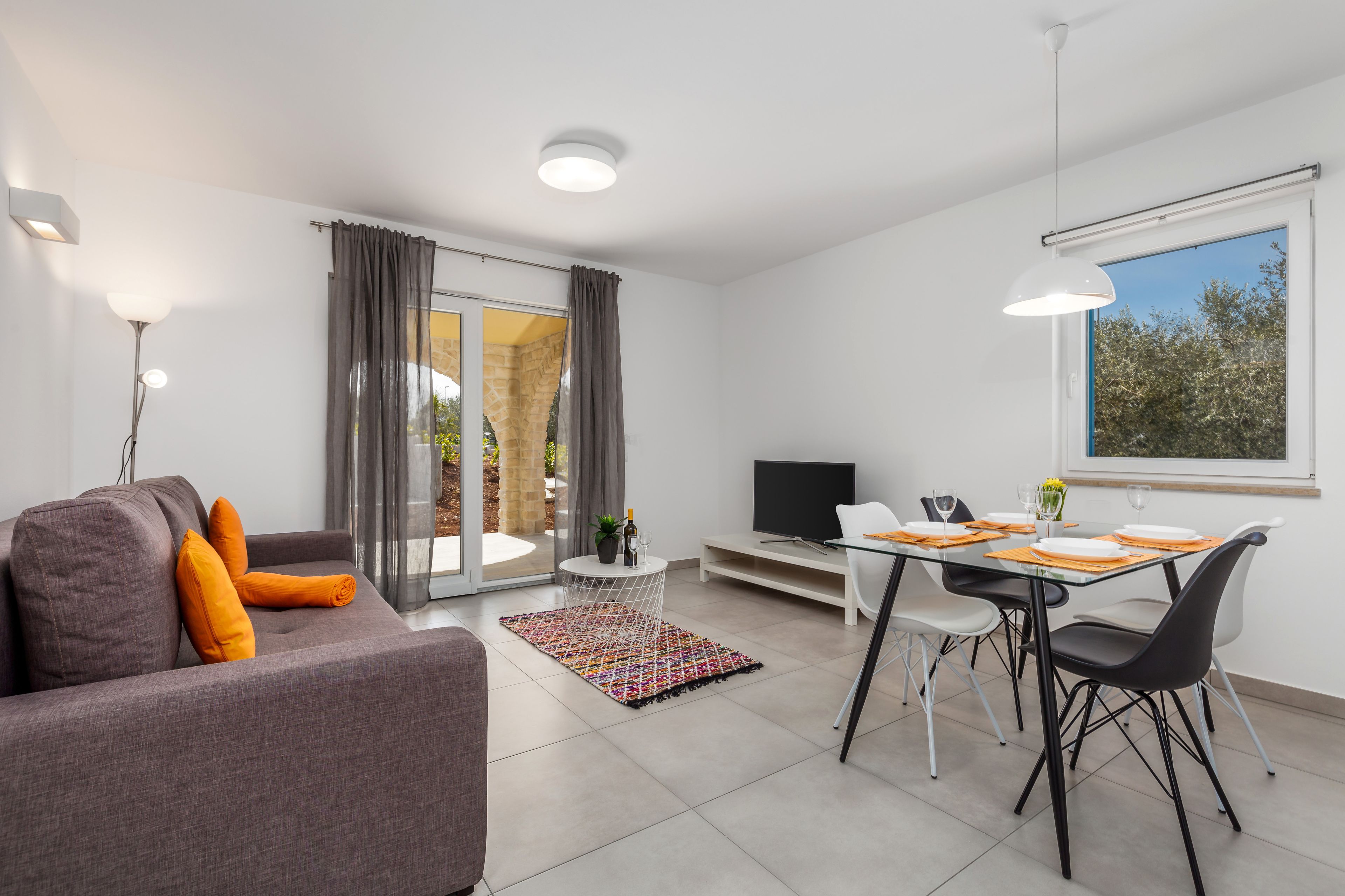 Una sala de estar moderna con un sofá gris, una mesa de comedor con sillas, un televisor y un ventanal con vistas a árboles.