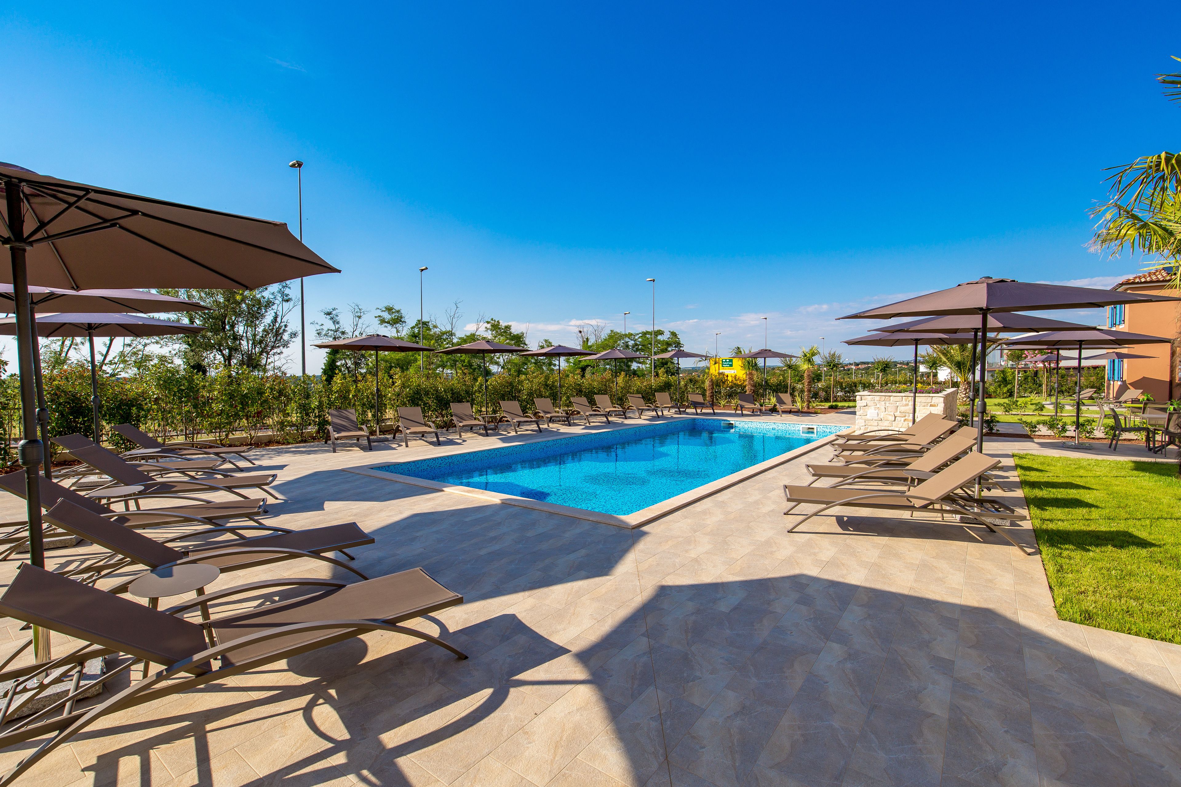 Une piscine entourée de verdure, avec des chaises longues et des parasols, sous un ciel dégagé.
