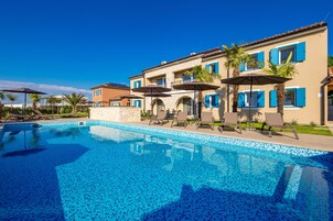 Una piscina con sillas de descanso y sombrillas, rodeada por un edificio con contraventanas azules y tejado de baldosas.