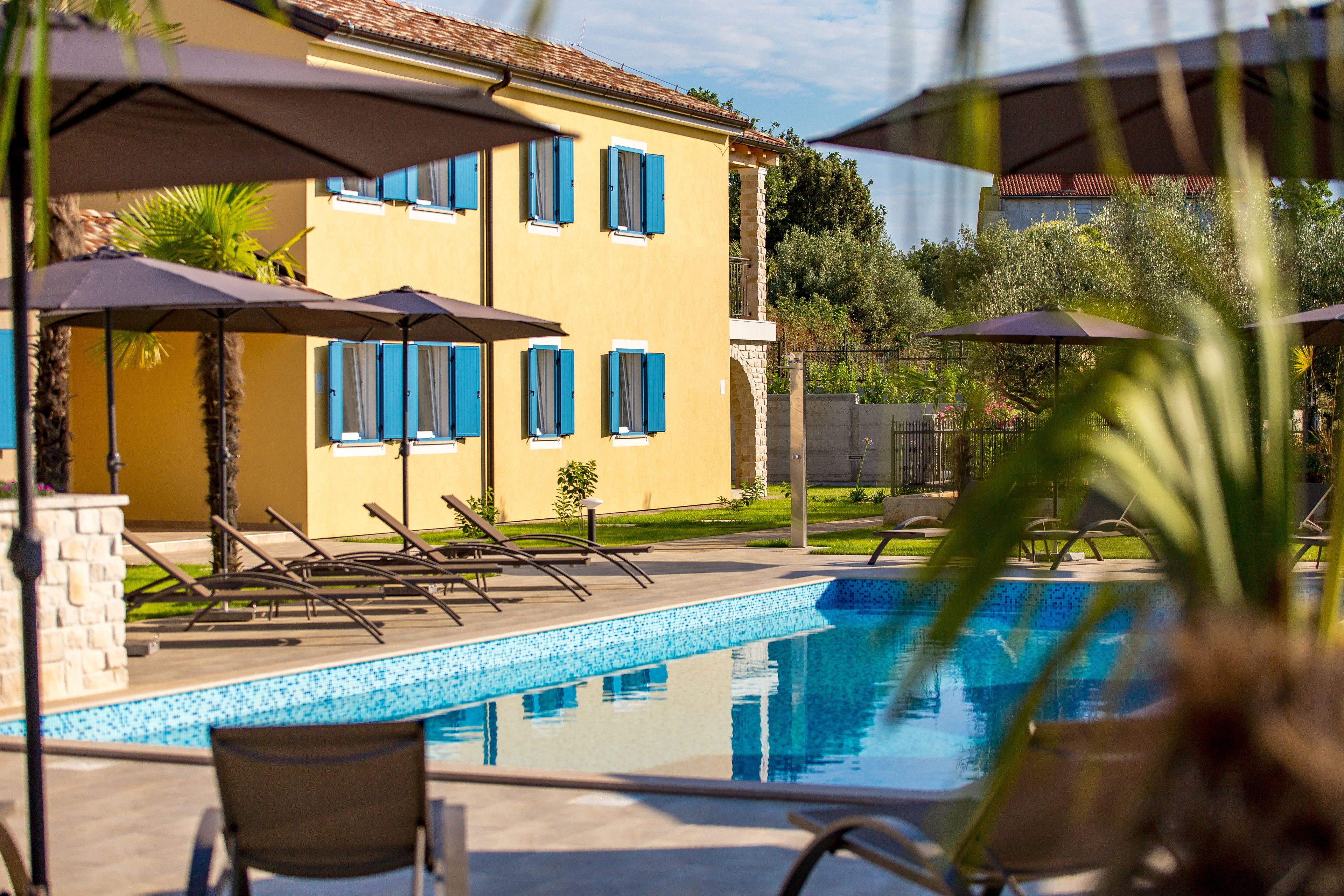 A pool area with lounge chairs and umbrellas, a yellow building with blue shutters, and a clear blue pool.