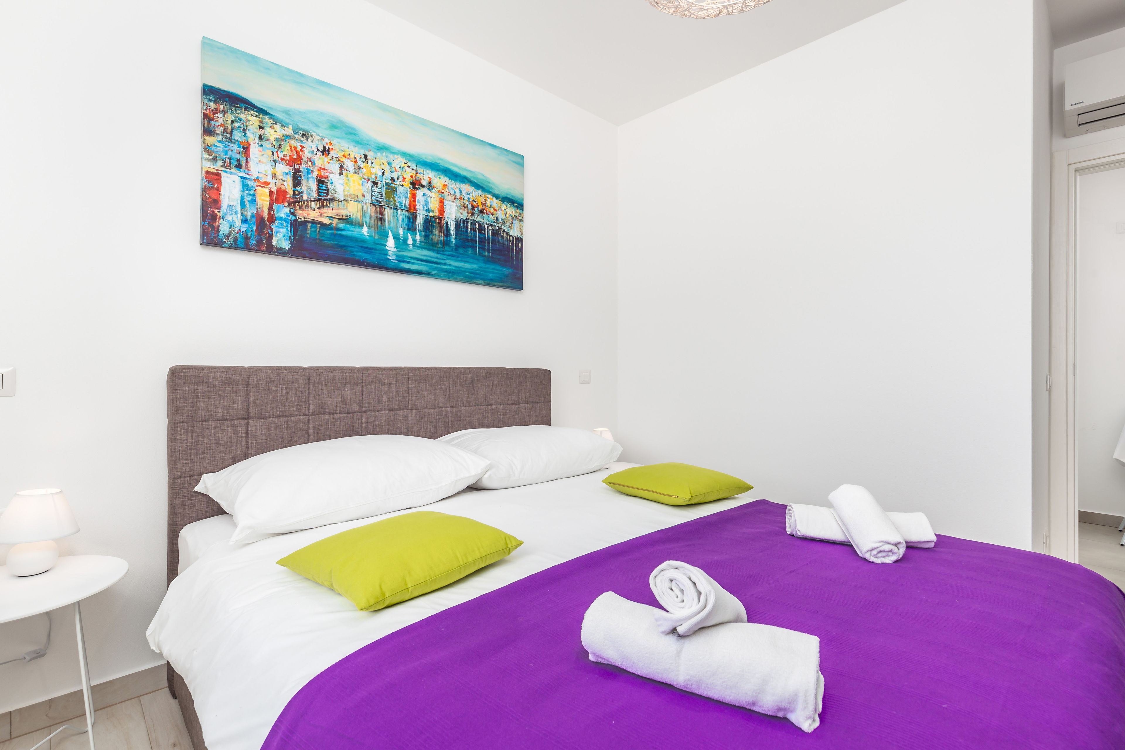 A modern hotel room with a large bed, white and green pillows, a purple bedspread, and a painting of a cityscape on the wall.