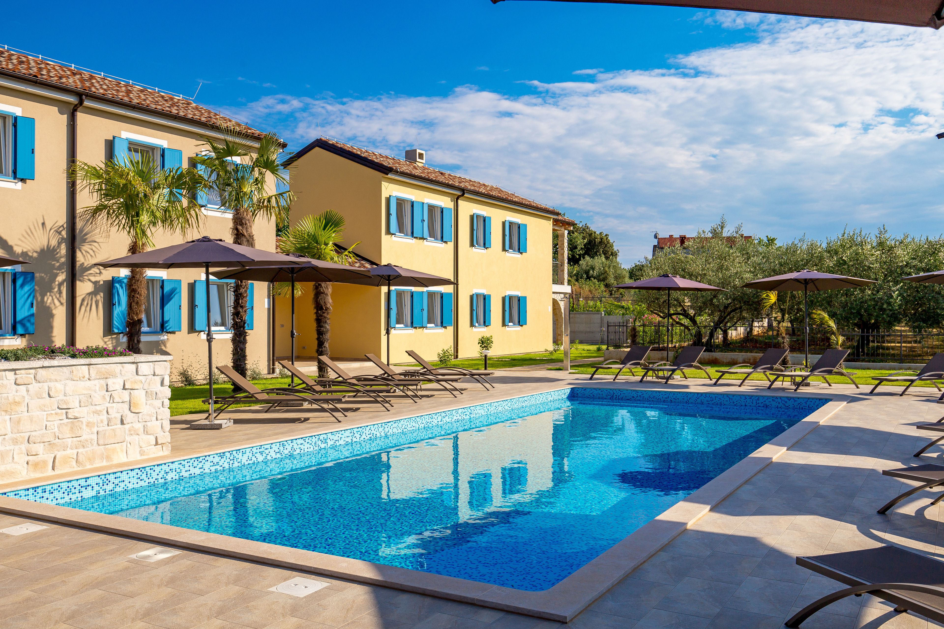 A swimming pool with lounge chairs and umbrellas, a stone wall, and a building with blue shutters and a tiled roof.