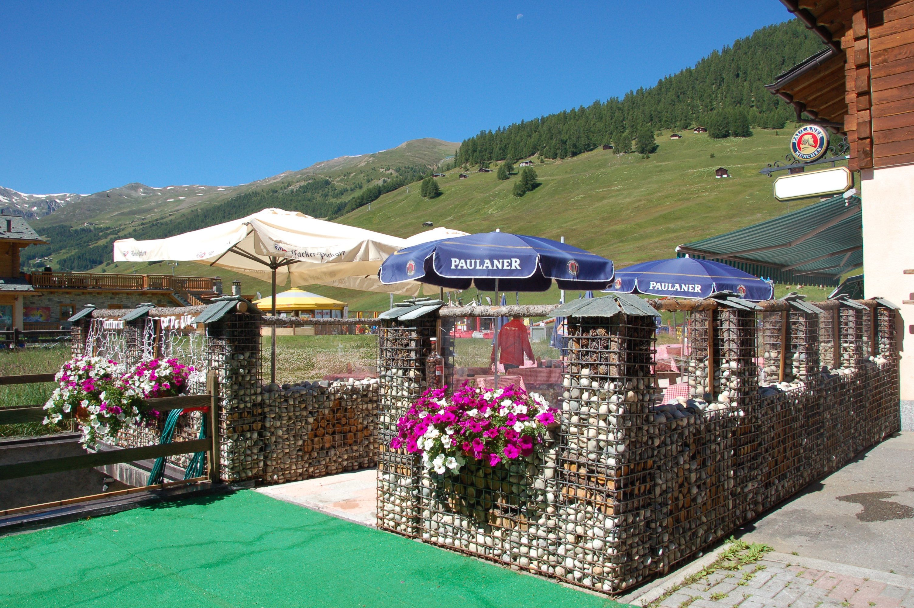 Terrassensitzgelegenheit mit PAULANER Sonnenschirmen, einer Steinwand mit Blumentöpfen und einem Bergpanorama im Hintergrund.